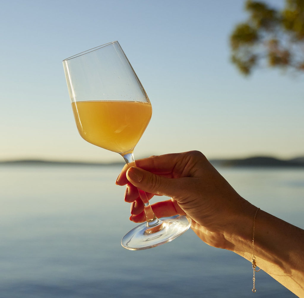Photo of a wine glass filled with pét-nat, against the setting sun over the ocean.